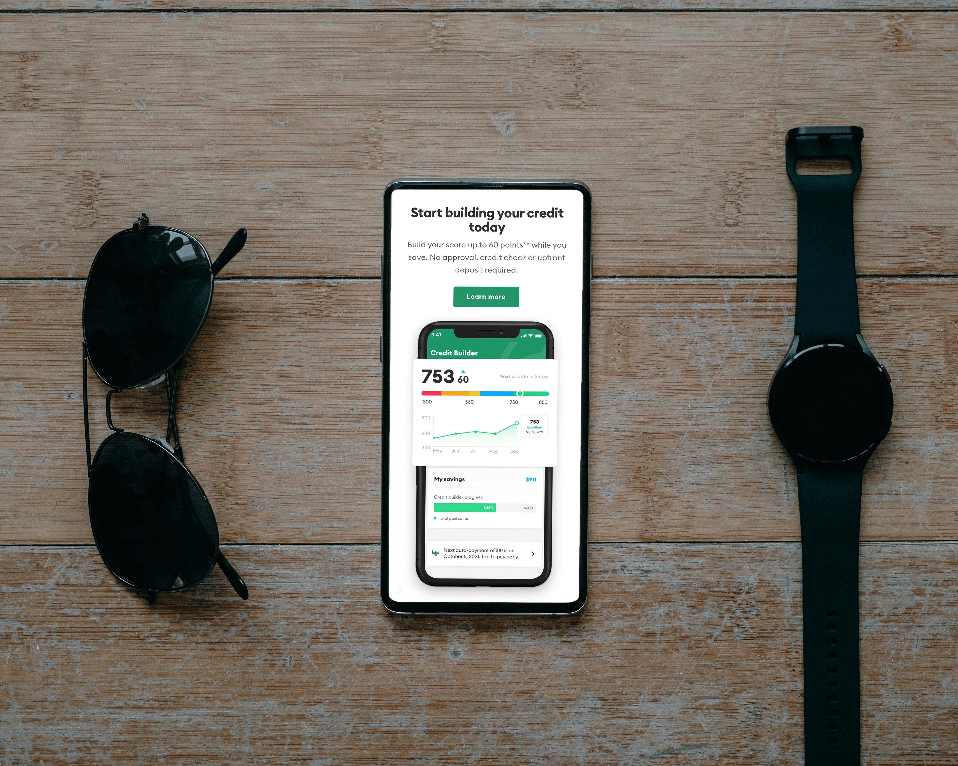 Smartphone showing credit builder app with score of 753, alongside sunglasses and watch on wood surface